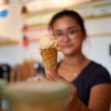 Eine junge Frau hält eine Eistüte mit zwei Kugeln Eis. Das Eis ist hellbeige und die Waffel hat ein Wabenmuster.