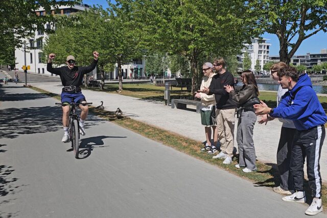 Ein junger Mann fährt auf dem Fahrrad, seine Freunde feuern ihn an.