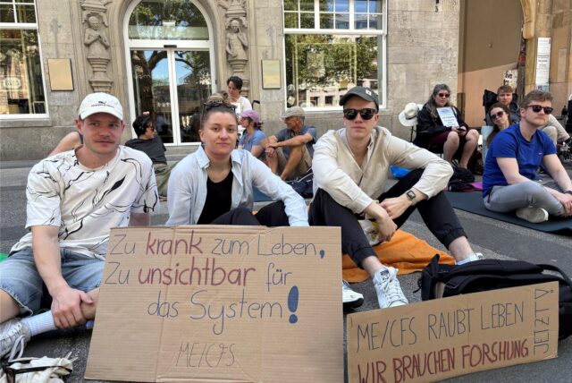 Etwa 100 Betroffene und Angehörige der Multisystemerkrankung ME/CFS demonstrienden im Liegen vor der Reinoldikirche am Ostenhellweg in Dortmund, Eine Frau hält das Plakat hoch mit der Aufschrift: „Wir fordern Inklusion und Forschung ME/CFS“.