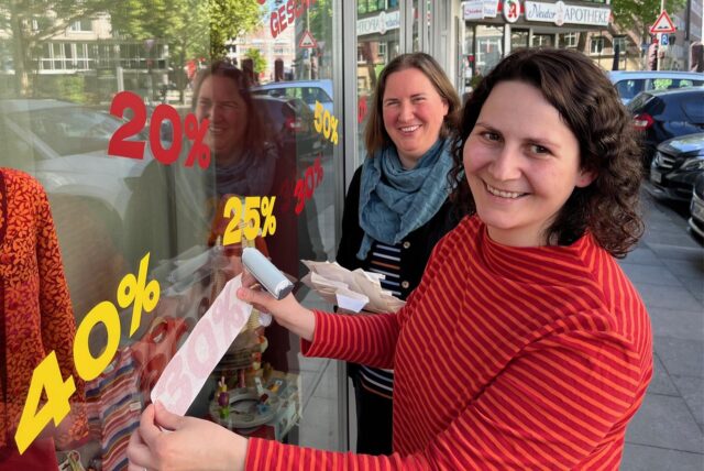 Zwei Frauen bekleben das Schaufenster eines Geschäfts mit Hinweisen auf Sonderangebote.