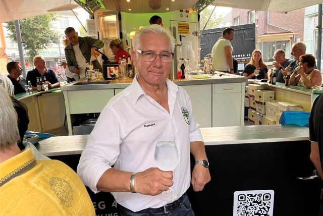 Gerhard Könecke steht beim Feierabendmarkt in Herten-Langenbochum vor einem Getränkestand und hält ein Glas Wasser in der Hand.