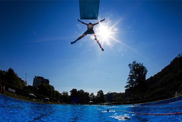 Ein Junge springt bei Sonnenschein und von einem Sprungturm in das Schwimmbecken eines Freibades.