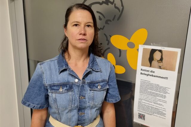 Eine Frau steht neben einem Plakat, auf dem „Rettet die Beleghebammen“ steht.