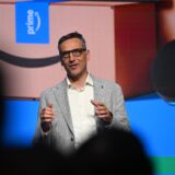 Der Amazon-Deutschlandchef, Rocco Bräuniger, steht bei einer Präsentation von Amazon in Dortmund  auf einer Bühne vor einer Leinwand und spricht zu Journalisten und Mitarbeitenden.