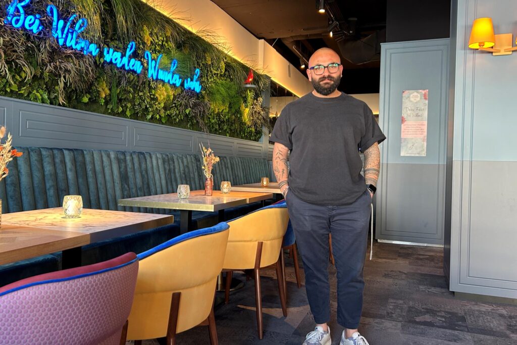 Can Korkmaz steht in seinem Restaurant Wilma Wunder. Im Hintergrund sind Tische und Stühle, eine grüne Wand und eine Neonreklame zu sehen, Can Korkmaz steht in seinem Restaurant Wilma Wunder. Im Hintergrund sind Tische und Stühle, eine grüne Wand und eine Neonreklame zu sehen,