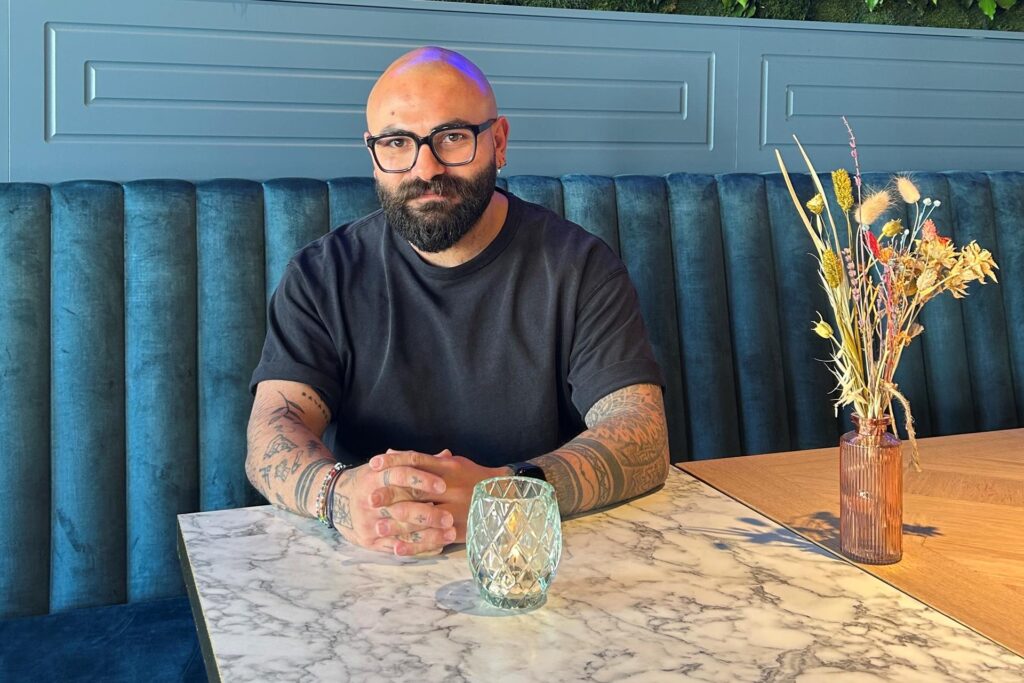 Can Korkmaz sitzt mit Brille und Tattoos an einem Tisch in der Wilma Wunder. An der Wand ist ein grünes Pflanzenarrangement mit blauer Neon-Aufschrift zu sehen. Can Korkmaz sitzt mit Brille und Tattoos an einem Tisch in der Wilma Wunder. An der Wand ist ein grünes Pflanzenarrangement mit blauer Neon-Aufschrift zu sehen.