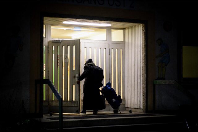 Eine Person in Mantel zieht ihr Hab und Gut in einem Rollkoffer hinter sich her und betritt in der Dunkelheit eine erleuchtete Unterkunft.