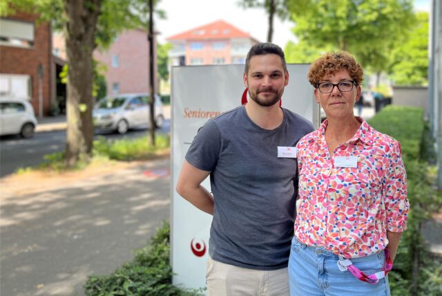Zwei Personen posieren vor einem Schild mit der Aufschrift „Senioren“. Beide Personen tragen Namensschilder.