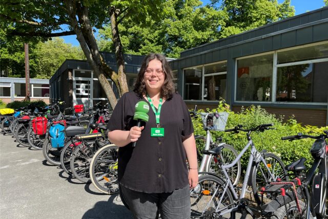 MZ-Volontärin Laura Wunderlich berichtet live vom Pedelec-Training für Senioren in Marl und steht vor dem Start der Übertragung mit dem Mikro in der Hand vor dem Fahrradständer mit Fahrrädern an der insel-VHS in Marl.