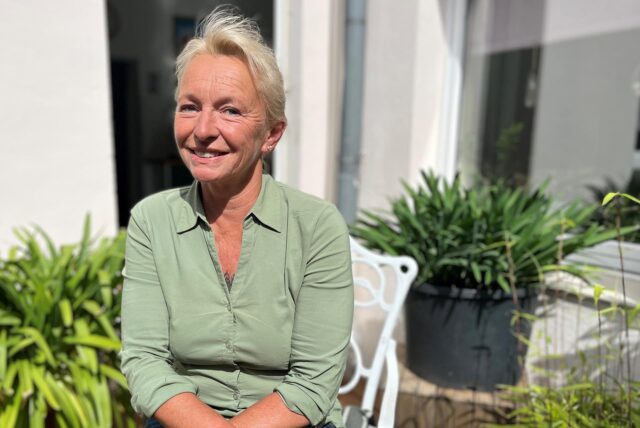 Petra Kussler sitzt in ihrem Hof in der Sonne und lächelt in die Kamera.