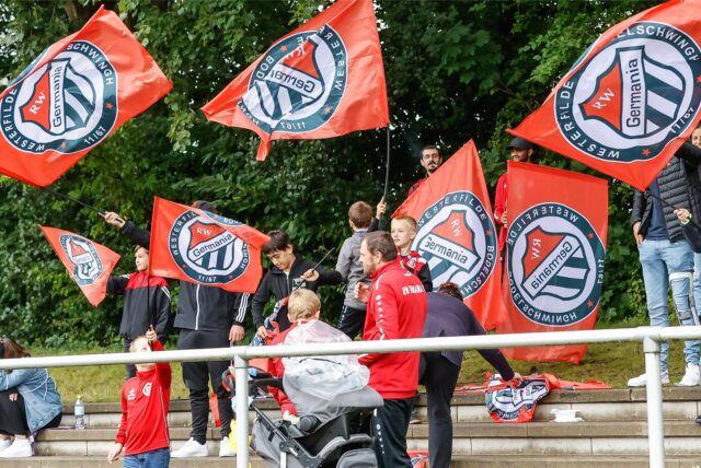 Fans von Rot Weiß Germania mit Vereinsfahnen auf den Tribünenstufen des Sportplatzes Im Odemsloh.