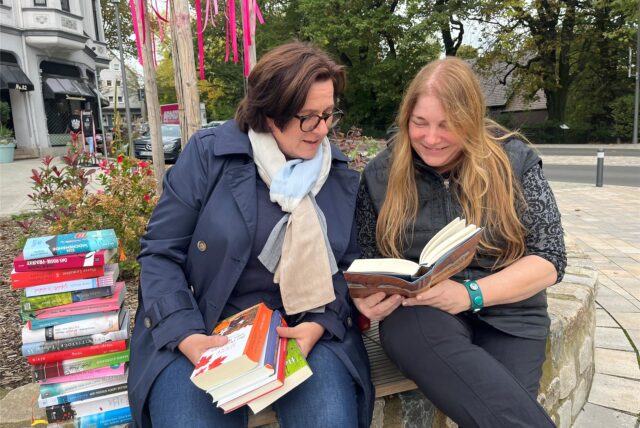 Zwei Frauen aus Recklinghausen halten Bücher in den Händen. Sie sitzen auf einer Mauer, im Hintergrund ist ein Beet zu sehen.