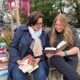 Zwei Frauen aus Recklinghausen halten Bücher in den Händen. Sie sitzen auf einer Mauer, im Hintergrund ist ein Beet zu sehen.