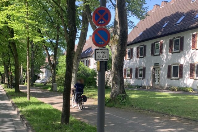 Ein absolutes Halteverbotsschild, ein eingeschränktes Halteverbotsschild und eine zeitliche Eingrenzung hängen an einem Mast. Auf einem Radweg fährt eine Radfahrerin.