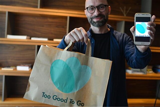 Ein Mann präsentiert eine Papiertüte und ein Smartphone mit dem Logo der App „Too Good To Go“. Die App zielt darauf ab, Lebensmittelverschwendung zu reduzieren, indem sie es Restaurants und Geschäften ermöglicht, überschüssige Lebensmittel zu reduzierten Preisen anzubieten.