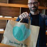 Ein Mann präsentiert eine Papiertüte und ein Smartphone mit dem Logo der App „Too Good To Go“. Die App zielt darauf ab, Lebensmittelverschwendung zu reduzieren, indem sie es Restaurants und Geschäften ermöglicht, überschüssige Lebensmittel zu reduzierten Preisen anzubieten.