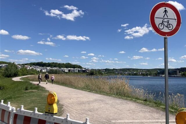 Ein Schild, das Fußgängern und Radfahrern den Weg verbietet, steht an einem Weg entlang eines Sees unter einem blauem Himmel mit Wolken. Im Hintergrund sind Häuser und ein bewaldeter Hügel zu sehen, während Spaziergänger den Weg nutzen.