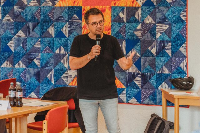 Ein Mann hält ein Mikrofon und gestikuliert vor einem Publikum während einer Präsentation. Im Hintergrund ist eine Patchworkdecke mit blauen und orangefarbenen Mustern zu sehen, während die Zuhörer an Tischen sitzen.