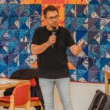Ein Mann hält ein Mikrofon und gestikuliert vor einem Publikum während einer Präsentation. Im Hintergrund ist eine Patchworkdecke mit blauen und orangefarbenen Mustern zu sehen, während die Zuhörer an Tischen sitzen.