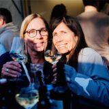 Zwei Besucherinnen des Feierabendmarktes prosten mit ihren Weingläsern der Kamera zu.