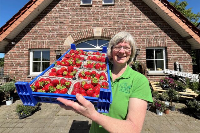 Eine Frau mit einer Transportbox voller Erdbeeren vor einem Hofladen.