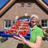 Eine Frau mit einer Transportbox voller Erdbeeren vor einem Hofladen.