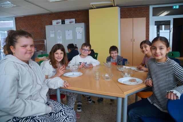 Sechs Kinder sitzen an einem Frühstückstisch und schauen in die Kamera.