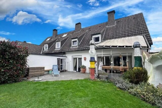 Die Doppelhaushälfte verfügt über einen Garten mit Wiese und Beet sowie einer Terrasse auf der ein gestreifter Liegestuhl steht.