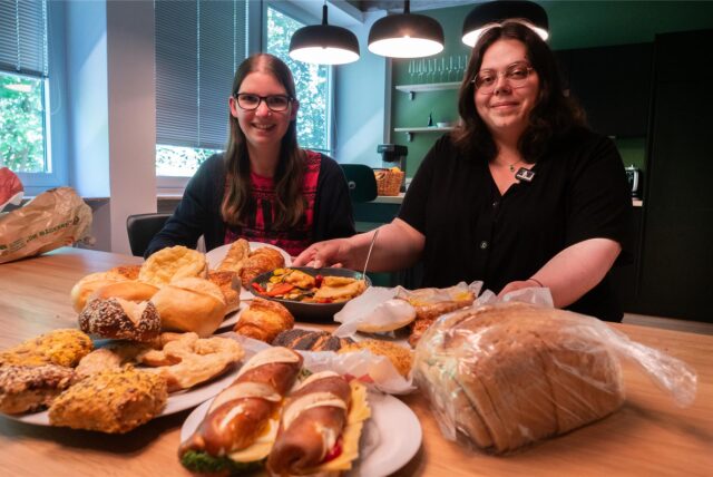 Katharina Göke (links) und Laura Wunderlich sitzen am Küchentisch der MZ-Redaktion. Vor ihnen stapeln sich Brötchen, Brot und andere Lebensmittel.
