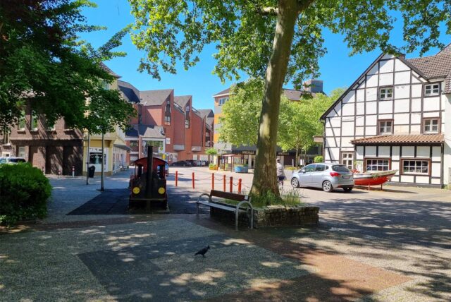 Eine Ansicht eines städtischen Platzes mit einer Spielplatz-Dampflokomotive und einer Bank im Vordergrund. Im Hintergrund sind mehrere Gebäude unterschiedlicher Bauart zu sehen, darunter ein Fachwerkhaus.