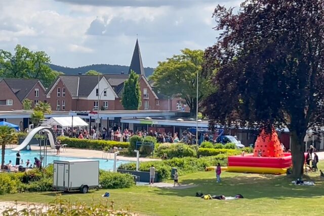 Freibad mit vielen Menschen und aufgebauten Zelten sowie Verkaufsständen und einer Hüpfburg. Grüne Wiese im Vordergrund