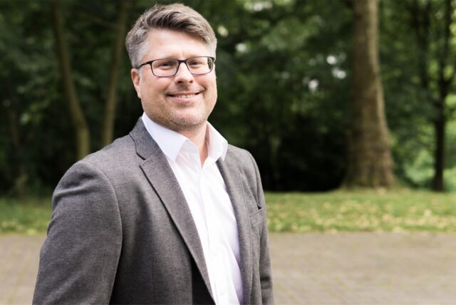Ein Mann mit Brille lächelt in die Kamera. Er trägt ein graues Jackett und ein weißes Hemd, während er vor einem unscharfen Hintergrund von Bäumen steht.