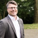 Ein Mann mit Brille lächelt in die Kamera. Er trägt ein graues Jackett und ein weißes Hemd, während er vor einem unscharfen Hintergrund von Bäumen steht.