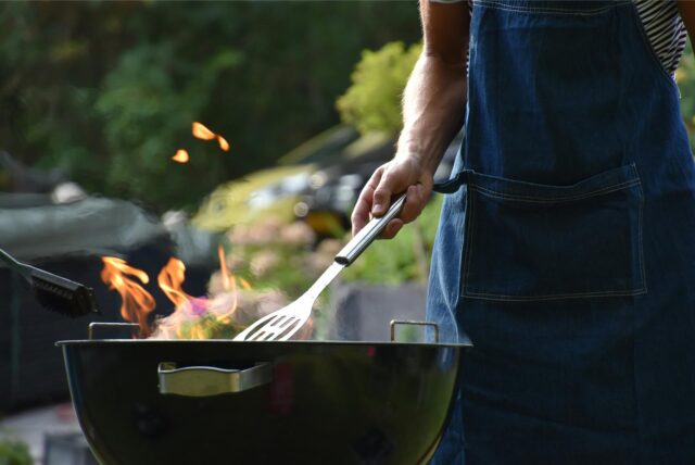 Mann in Kochschürze und mit Pfannenwender grillt an einem runden Standgrill