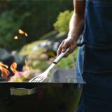 Mann in Kochschürze und mit Pfannenwender grillt an einem runden Standgrill