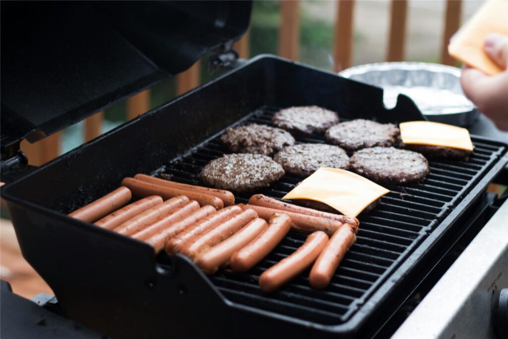 Auf einem Grillrost werden zahlreiche Würstchen, fünf Burgerpattys und zwei Scheiben Käse gegrillt Auf einem Grillrost werden zahlreiche Würstchen, fünf Burgerpattys und zwei Scheiben Käse gegrillt
