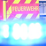 Ein Fahrzeug der Feuerwehr steht bei einem Einsatz auf einer Straße, während im Vordergrund das Blaulicht eines Polizeifahrzeugs zu sehen ist. (Symbolbild)