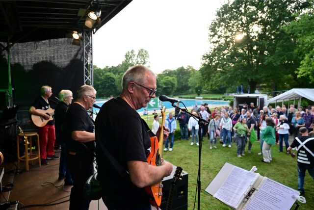 Eine Band macht Musik auf einer Bühne, im Hintergrund das Publikum und ein Freibad.