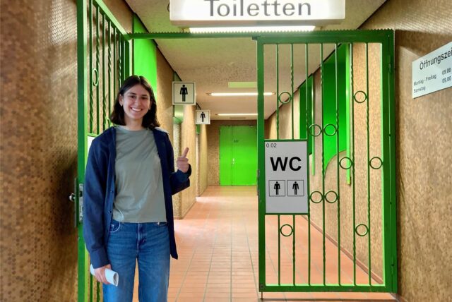 Xenia zeigt Daumen vor dem Eingang zur Toilettenanlage Kampstraße in Dortmund den Daumen hoch.