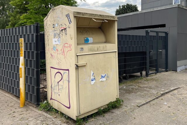 Eine öffentliche Altkleidersammlung steht auf einem Bürgersteig neben einem Zaun. Der Behälter ist mit Graffiti und Aufklebern versehen.