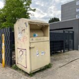 Eine öffentliche Altkleidersammlung steht auf einem Bürgersteig neben einem Zaun. Der Behälter ist mit Graffiti und Aufklebern versehen.