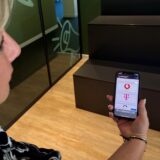 Vodafone-Kundin blickt auf ihren Handybildschirm mit den Firmenlogos von Vodafone, Telekom und O2.