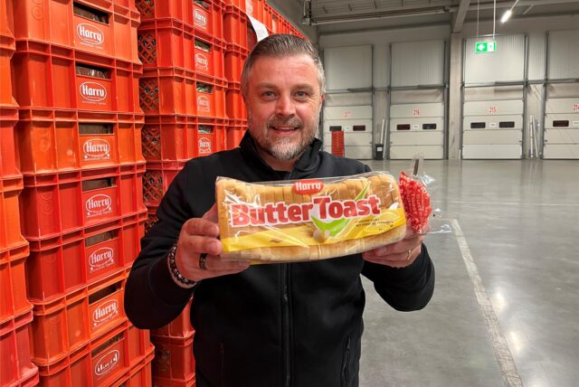 Ein Mann präsentiert eine Packung Butter Toast der Marke Harry vor einem Stapel leerer Brotkisten derselben Marke. Im Hintergrund sind mehrere Rolltore mit Nummern zu sehen.