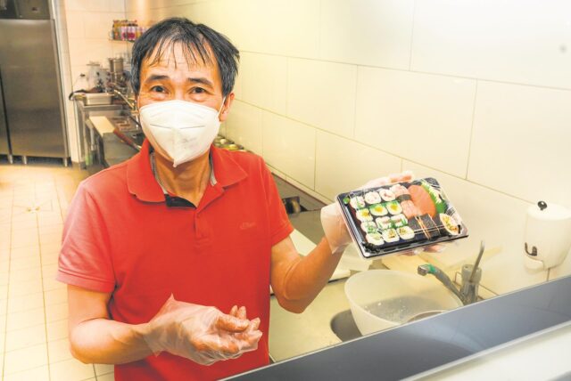 Ein Mann steht hinter einer Theke und hält einen Behälter mit einer Portion Sushi in der Hand.