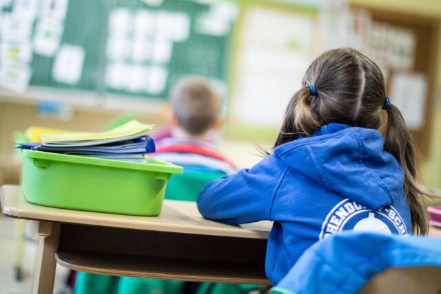 Eine Schülerin sitzt mit dem Rücken zur Kamera an einem Schreibtisch in einem Klassenzimmer. Im Hintergrund ist eine Lehrerin an der Tafel zu sehen.