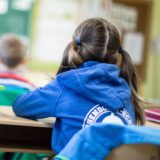 Eine Schülerin sitzt mit dem Rücken zur Kamera an einem Schreibtisch in einem Klassenzimmer. Im Hintergrund ist eine Lehrerin an der Tafel zu sehen.