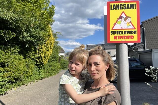 Sophie De Manos, Anwohnerin der Ulrichstraße in Datteln-Horneburg, mit ihrer Tochter Yuna auf dem Arm. Daneben hängt ein Schild: „Langsam! Spielende Kinder“.