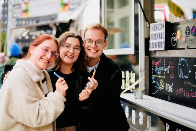 Drei Menschen stehen vor einem Foodtruck und lächeln.