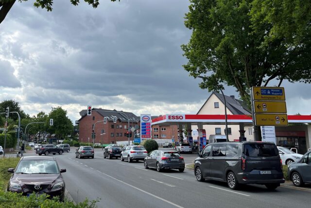Auf dem Foto ist eine Esso-Tankstelle an einer belebten Straße zu sehen. Mehrere Autos warten an einer Ampel, während im Hintergrund der Himmel bewölkt ist.