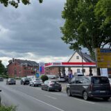 Auf dem Foto ist eine Esso-Tankstelle an einer belebten Straße zu sehen. Mehrere Autos warten an einer Ampel, während im Hintergrund der Himmel bewölkt ist.